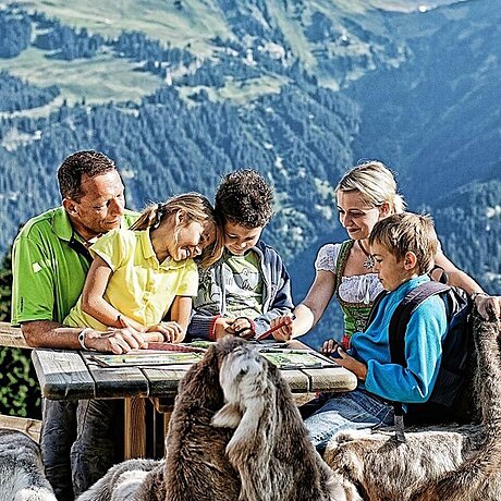 Eine Familie sitzt an einem Holztisch auf einer Alm und berät sich anhand einer Landschaftskarte