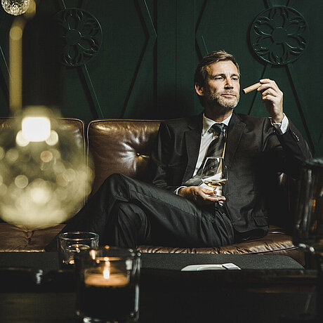 Ein Herr im Anzug genießt auf der Ledercouch der stimmungsvollen Smokers Lounge im Löwen Hotel Montafon eine gute Zigarre und ein Glas Wein