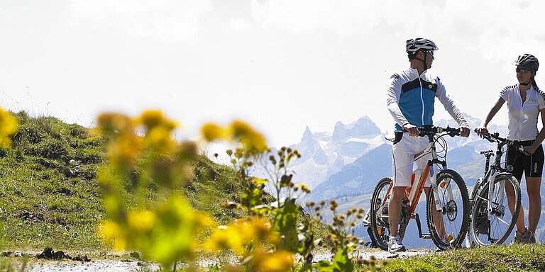 Zwei Mountainbiker stehen auf einem Feldweg und unterhalten sich vor dem beeindruckenden Bergpanorama