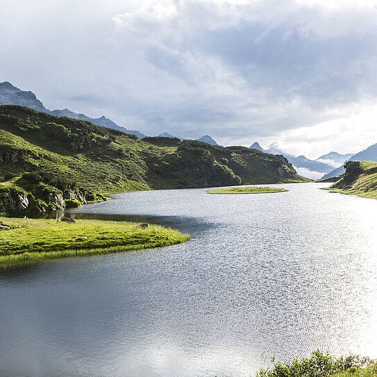 Der Langsee zieht sich durch grüne Hügel. Am Himmel hängen einige Wolken.