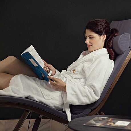Eine Frau im Bademantel sitzt auf einer Liege und liest ein Magazin