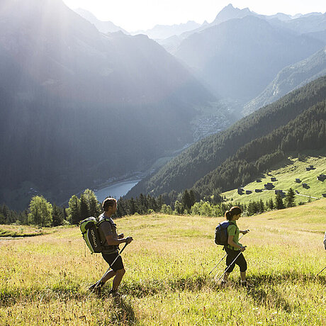 Zwei Wanderer folgen einem Pfad durch das hohe Gras eines Berghangs. Im Tal liegt ein See.