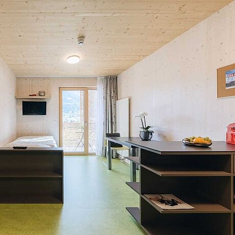 Das Einzelzimmer im Teamhaus des Löwen Hotel Montafon mit einem Einzelbett, einem Esstisch mit Stuhl und einem großen Fenster mit Blick auf die Berge.